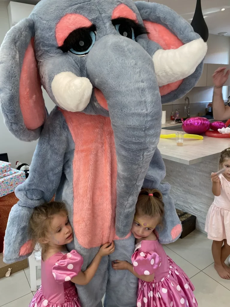 Danny le Magicien propose son Animation de Peluche Géante pour l'Anniversaire d'Enfants. "Calinette" l'éléphant rose est embrassée par deux adorables petites filles habillées également en rose.